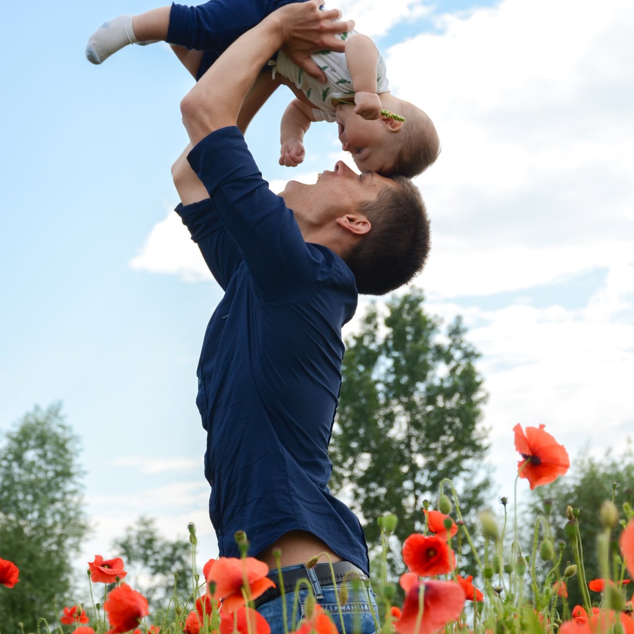 Vater hebt sein Kind hoch, er steht in einem Feld mit Mohnblumen.