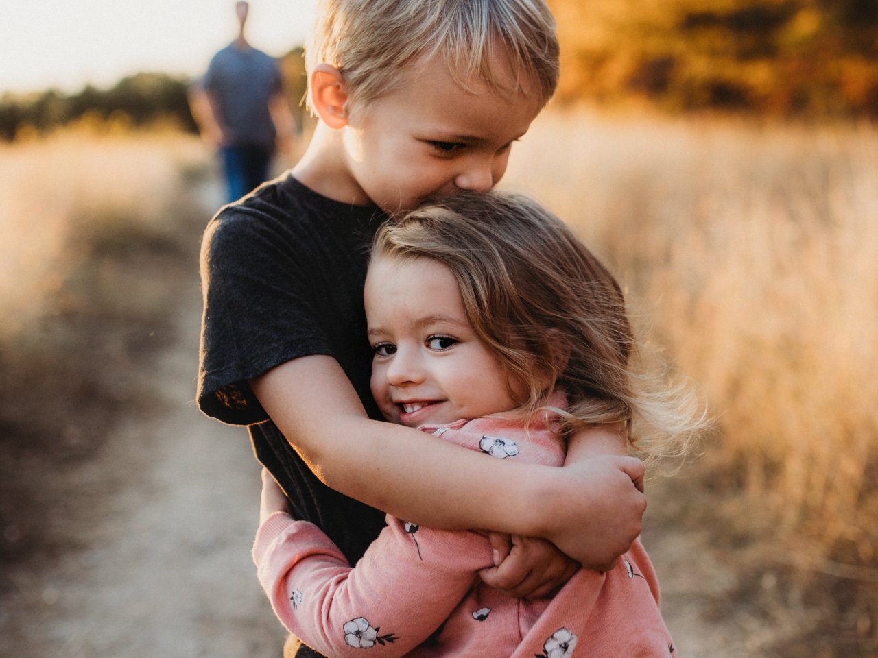 Junge umarmt seine kleine Schwester, im Hintergrund sieht man die Eltern. 